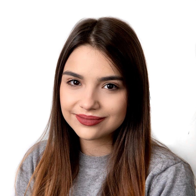 A person soft smiling in front of an all white background, showing no teeth with long, straight brown hair that starts to turn ombre. They have on red matte lipstick and are wearing a grey sweater.