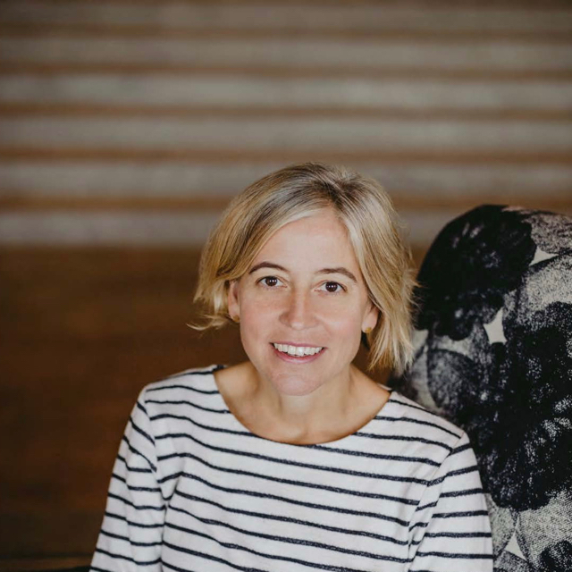 A person smiling in a chair. They are in a black and white striped long sleeve shirt and they have short dirty blonde hair. The chairs fabric print has black and white flowers on it.
