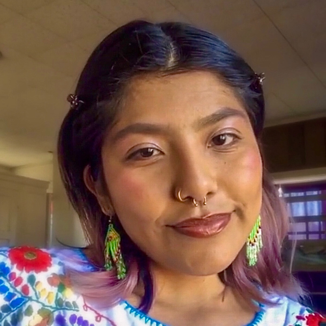 Person soft smiling in a room. They have short straight black hair with pink highlighted tips and are wearing colorful beaded earrings and a white cotton shirt with flowers on it.