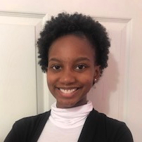 Person smiling in front of a white door. They have short curly black hair and are wearing a white turtle neck with a black blazer over it.
