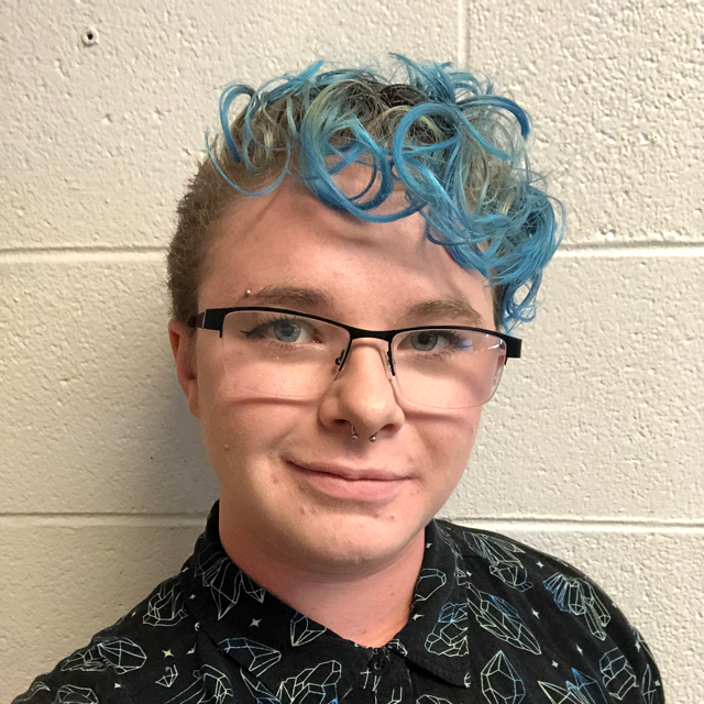 Person smiling in front of a white brick wall. They have short blonde curly blonde hair with blue tips and are wearing black glasses and a black button up with green and blue crystals on it.