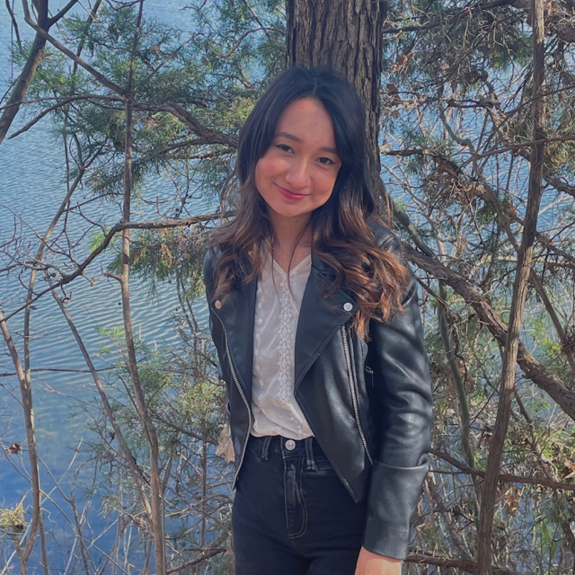 Person smiling in front of a tree with a body of water behind it. They have black hair with ombre highlights and are wearing a white shirt with a black leather jacket and pants.