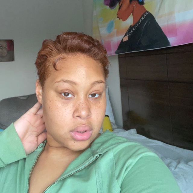 Person still-faced in a selfie pose in their bedroom. They have curly, short, auburn hair and are wearing a green zip up jacket.