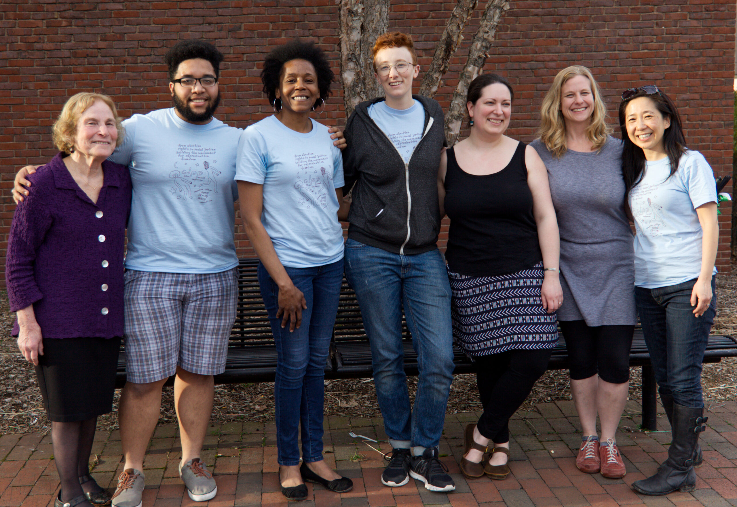 A group of people smiling in front of a bench, tree, and brick wall. They each have their arms around someone and are all showing their teeth. Some are wearing matching light blue t-shirts and others are wearing clothing like a black tank top, grey dress, or purple suit. Theres black curly and straight hair, blonde wavy hair, and short red hair present. Some are wearing boots, tennis shoes, flats, and gym shoes.