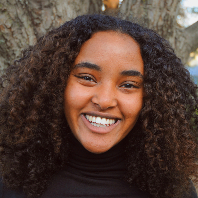 Person smiling in front of a tree. They have long dark brown curly hair and are wearing a black turtleneck.