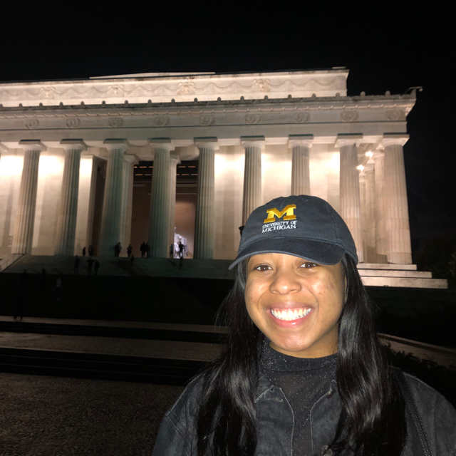Person smiling in front of a national monument. They have on a navy blue hair and grey shirt and jacket. They have long straight black hair.