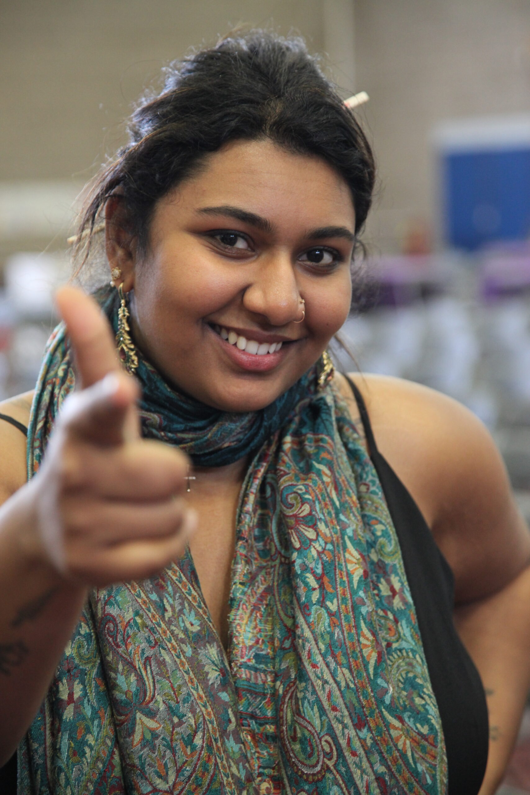 Person smiling with straight black hair in a bun, a nose piercing and turquoise patterned scarf. They are holding a finger gun sign up in a light-hearted matter.