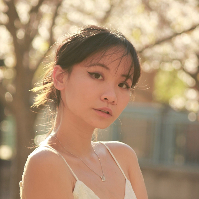 A person still-faced in front of a blurred nature background. They have straight black hair in a bun and bangs. They are wearing a necklace and white tank top as well.