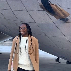 Person standing in front of the Chicago "Bean." They have long black braids, black glasses, and are wearing a white sweater, black pants, and a tan blazer.
